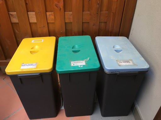 Three rectangular black bins, from left to right with a yellow, green, and blue lid, for separating waste.