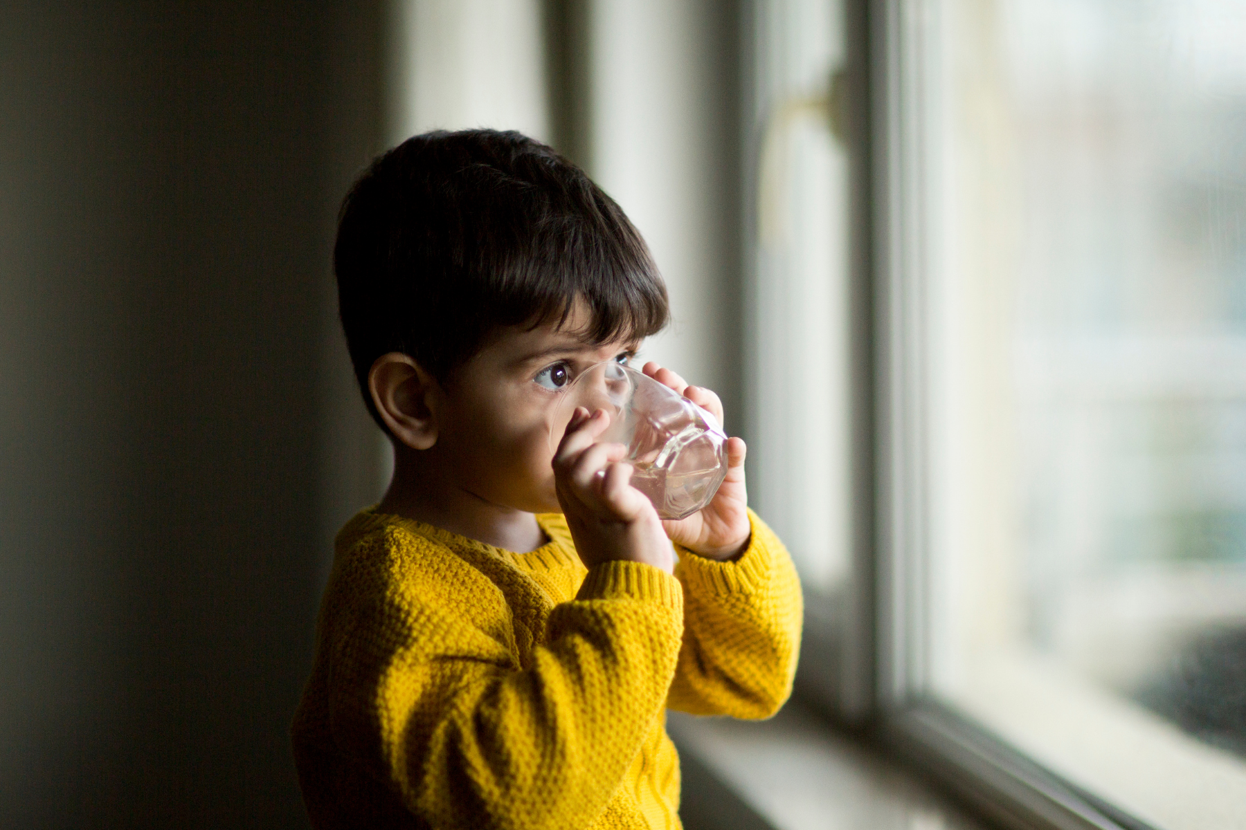 Child looking out of a window, drinking water from a glass.