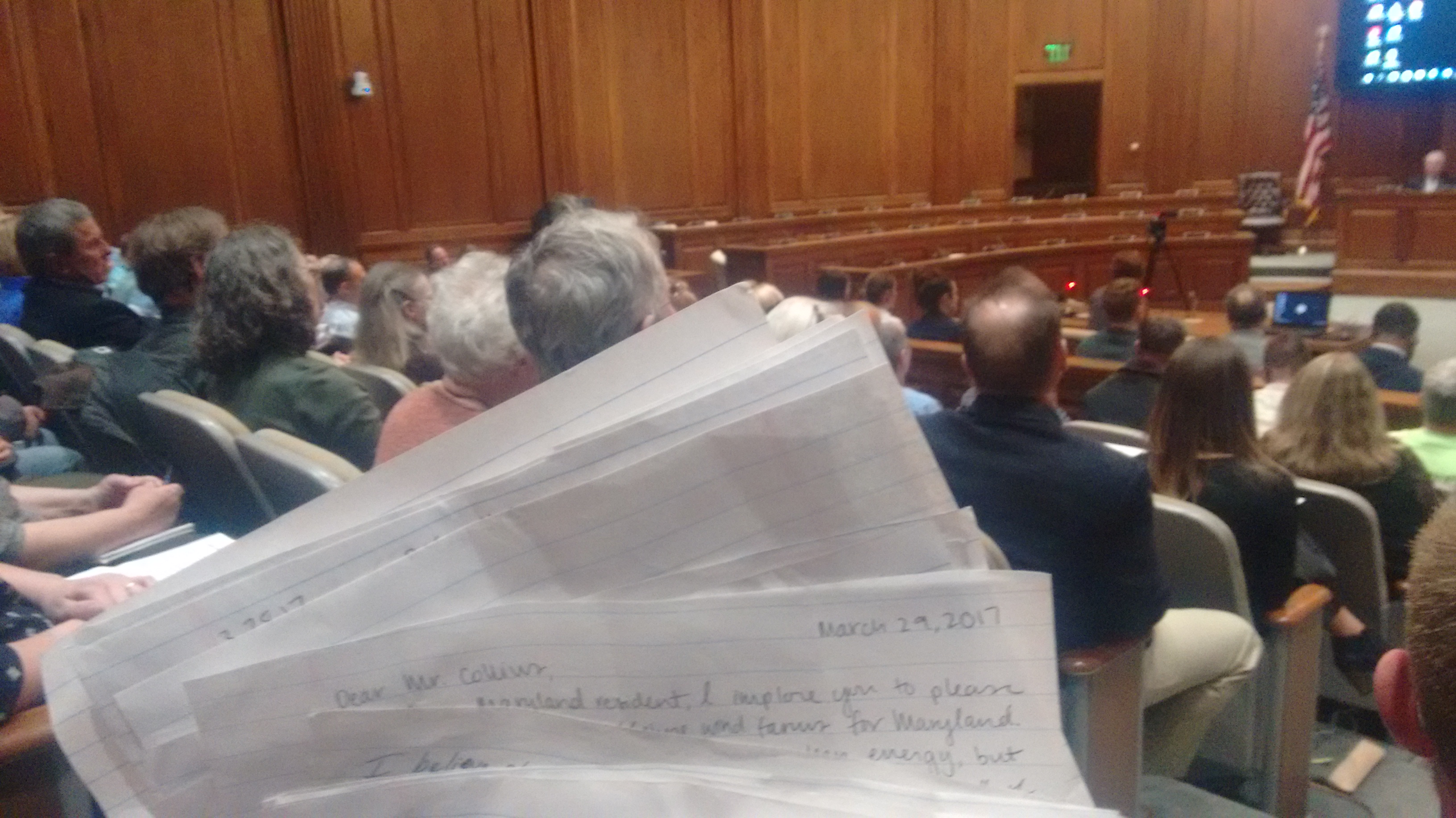 The public hearing room, full, with handwritten letters held in the foreground