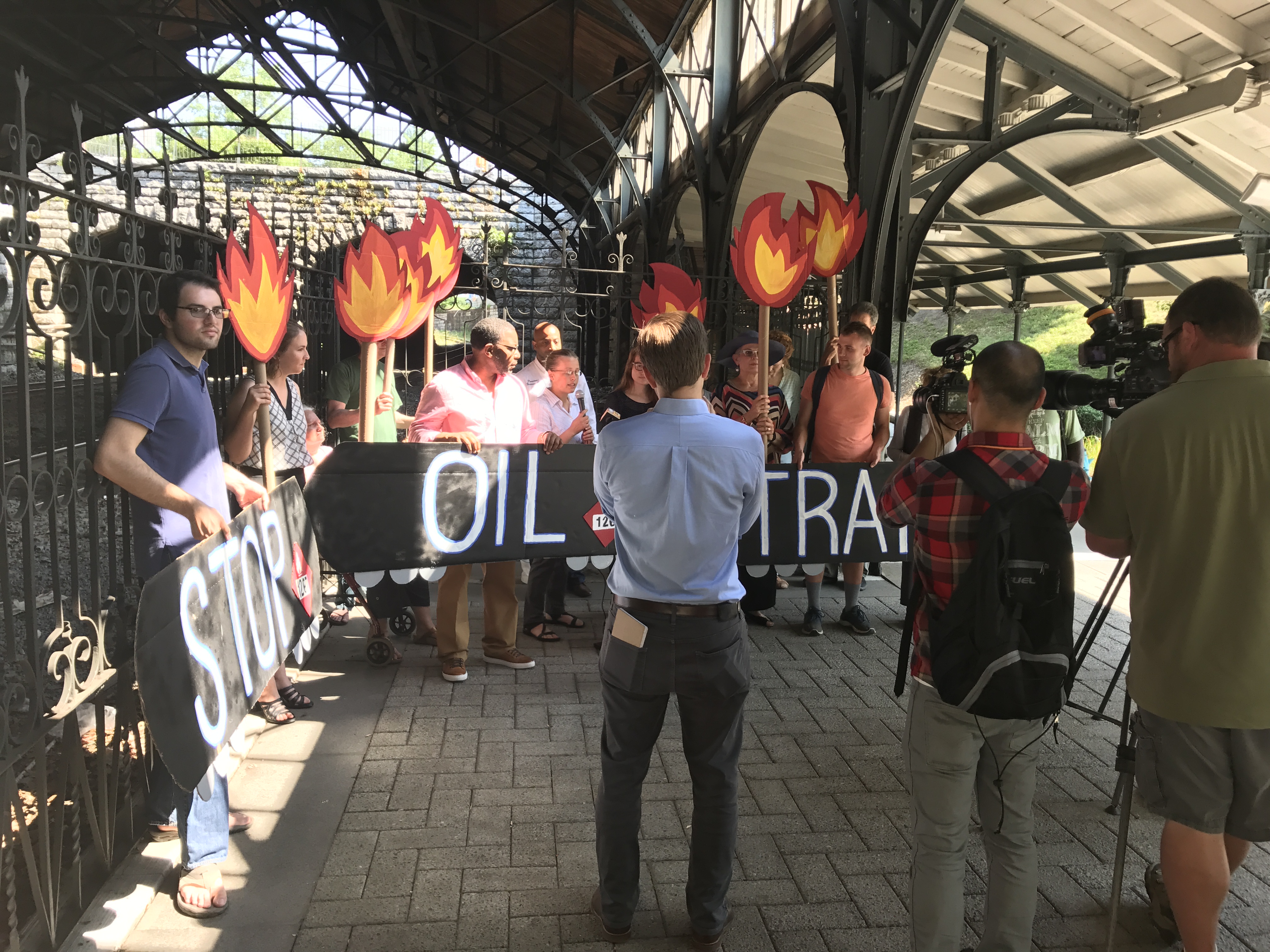 15 people hold signs saying "Stop Oil Trains" and showing train tank cars and flames as news media films